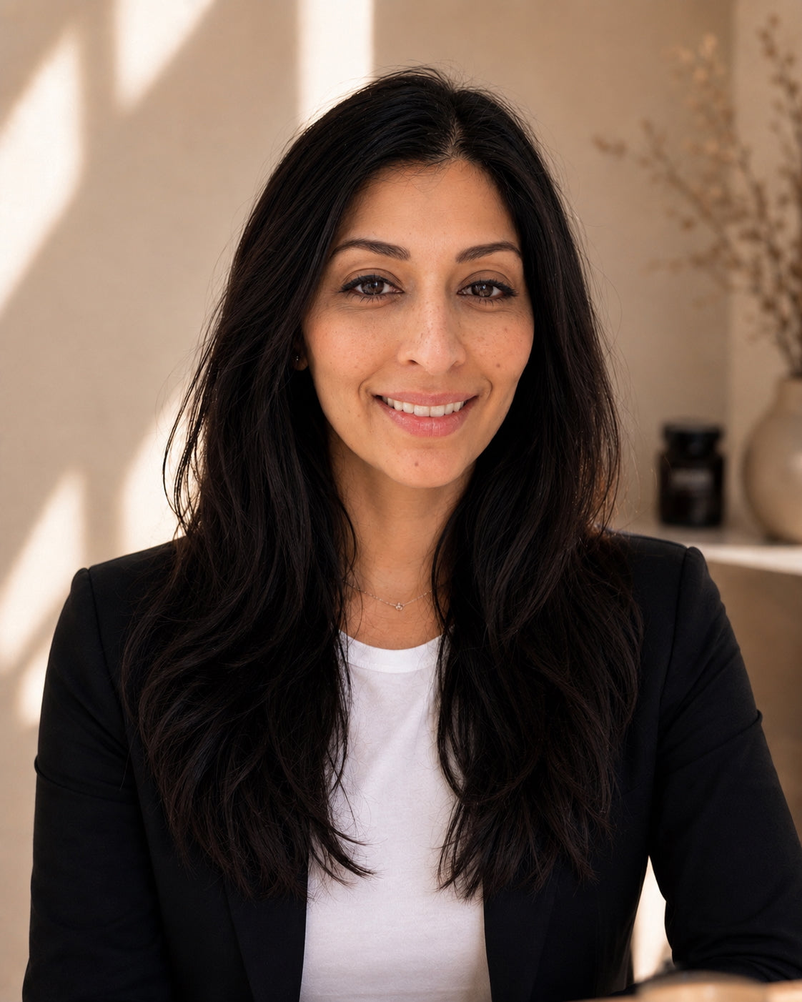 Woman with long dark hair wearing a black blazer over a white shirt, sitting indoors.